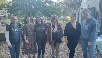 Six women are standing and smiling for the camera. The ground is woodchip and there are trees in the background.