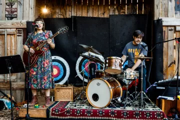 Two people perform on a small wooden stage outdoors. One is a woman wearing a floral dress playing guitar and singing. She is standing. The other person is a man seated and playing drums and singing.