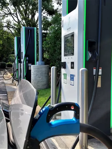 The hood of an electric car is shown with its charging port open and plugged into a charger in a parking lot surrounded by trees.