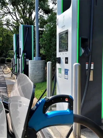 The hood of an electric car is shown with its charging port open and plugged into a charger in a parking lot surrounded by trees.