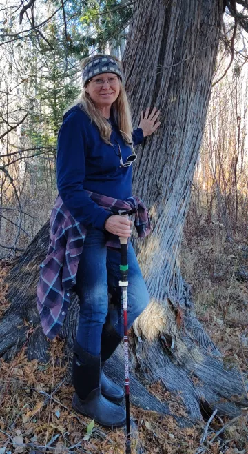 A woman with long blond hair a winter hat stands next to a tree with her arm outstretched so her hand is on the tree trunk. She is in a forest and is smiling.