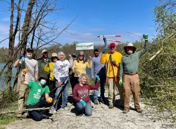 A group of people outside with various tools for invasive species removal. The sky is blue and there are trees in the background. One person is holding up a sign saying Weed Wrangle.