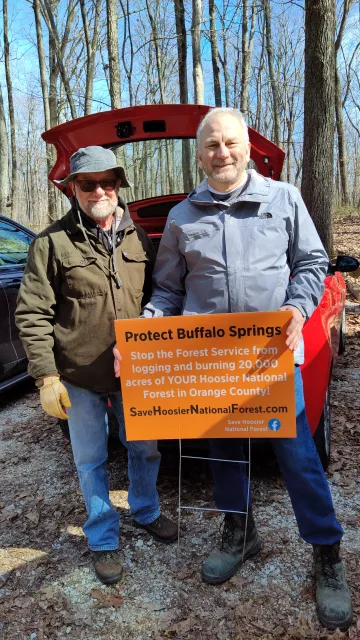 Two men stand in a parking lot with trees in the background. They are holding an orange sign which says Protect Buffalo Springs. Stop the Forest Service from logging and burning 20,000 acres of YOUR Hoosier National Forest in Orange County! SaveHoosierNationalForest.com
