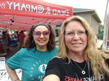A selfie of two women standing outside at an event. One has blonde hair and the other brown hair. They are both smiling. There is a red canopy set up in the background with the words Dead & Company in white.