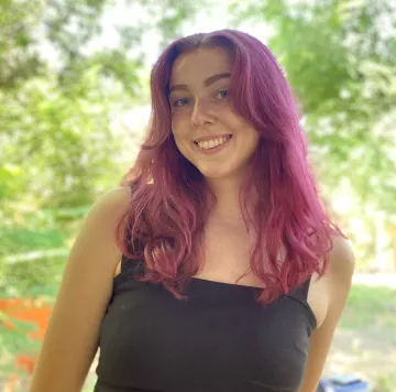 Amanda (white woman with plum colored hair) smiling in front of green foliage