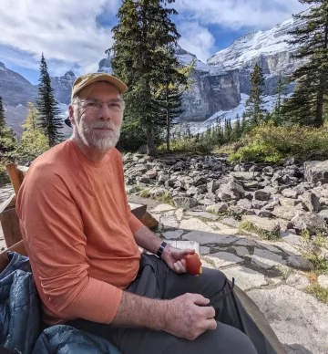 Photo of Jim Wylie eating an apple, sitting in a forest, surrounded by rocks, tall trees, and snow-capped mountains.