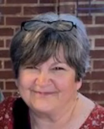 Debi is smiling at the camera with a brick background. She is wearing glasses on top of her head and a pink shirt.