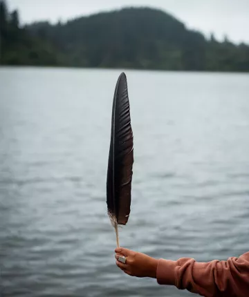 Condor feather being held by a hand wearing a silver ring with a lake in the background