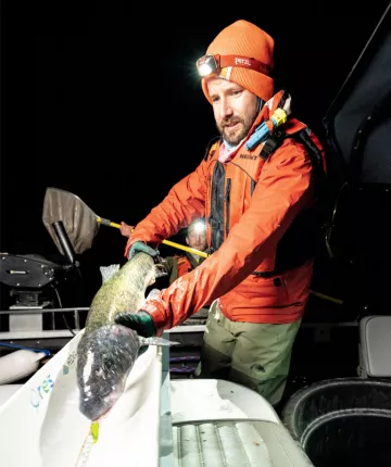 Steven Staiger wears an orange jacket and ski cap and holds out a large fish to be measured.
