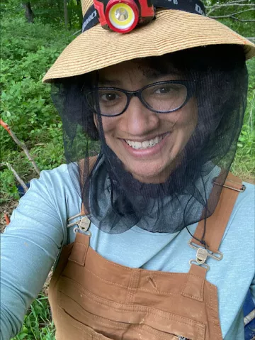 Tsion is smiling outdoors and wearing a hat with netting, a blue shirt, and brown overalls.