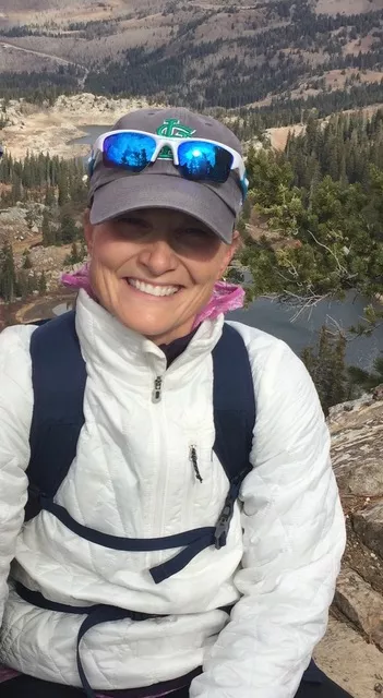 Helen is outdoors, with trees and mountains behind her. She is smiling at the camera. She is wearing a ball cap with sunglasses ont op, a white hiking jacket, and a book bag.