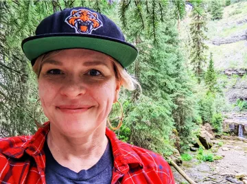 Katie is outdoors with pine trees and hills behind her. She is smiling at the camera wearing a navy hat with a tiger logo and green brim. She is also wearing a navy shirt with a red long sleeve over it.