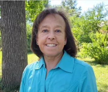 Stephanie is outdoors , with trees and bushes behind her. She is smiling at the camera and wearing a blue shirt.