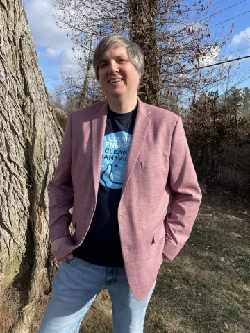A person stood wearing a pink jacket and blue pants. They are outdoors with grass and some trees behind them. They are smiling and look relaxed.