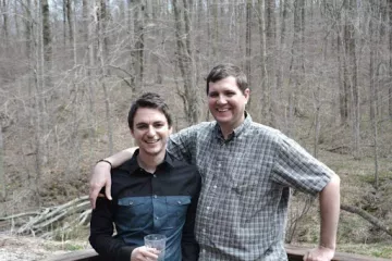 Two people stood with a forest backdrop. The taller person has their arm around the other. The trees are bare.