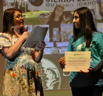 sherlyn on stage holding certificate while lisa looks at her and speaks into microphone