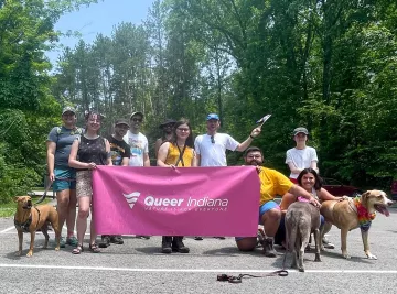 A group of people standing in a parking lot with trees behind them on a sunny day. There are a few dogs present too. A few of the people are holding up a pink banner saying Queer Indiana in white writing. They are smiling.