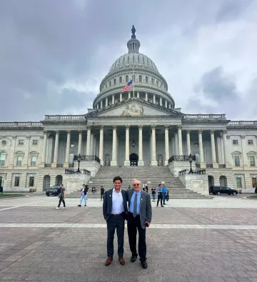 Two men stand in front of the Capitol Building in D.C. The building has a domed roof and several pillars at the front. The sky is cloudy.