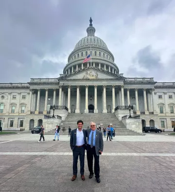 Two men stand in front of the Capitol Building in D.C. The building has a domed roof and several pillars at the front. The sky is cloudy.