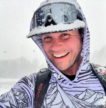 John Roden smiles wearing a snowy ski mask, hoodie, and backpack.