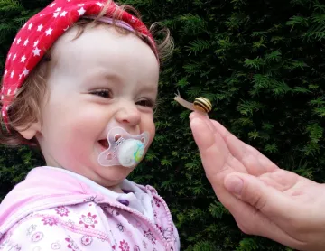 Photo of baby girl laughing as an adult hand holds a snail up to her