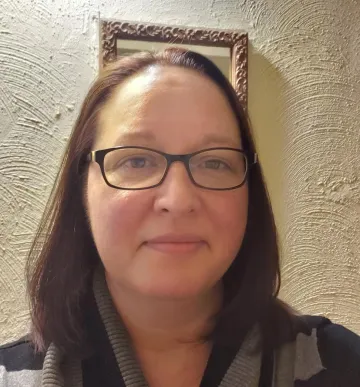 A head and shoulders shot of a woman with brown hair and dark glasses, indoors with an off-white wall behind her.