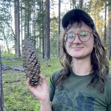 A portrait of Madeline Cowen standing in a forest holding a large pinecone