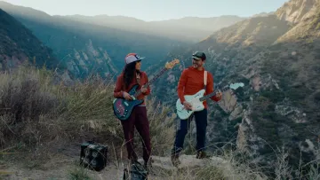 Picture of a woman and man playing electric guitar outdoors with a mountainscape behind them