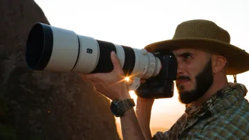 Picture of man with a beard and hat holding a large professional camera outdoors