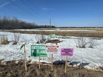 Signs at the edge of a field with some snow. Signs read “Entering Wetland”, “Buffer Zone: No Fueling or Maintenance” and “Invasive Management Area”