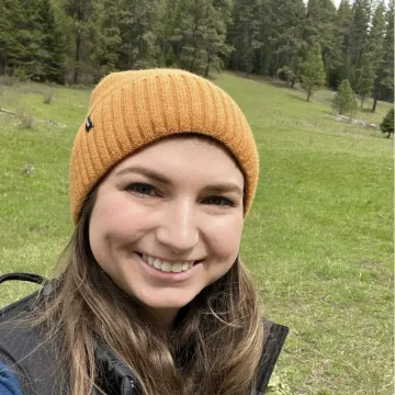 Krystal, a white woman with long hair and wearing a beanie, smiling at the camera while outdoors with trees and grass in the background