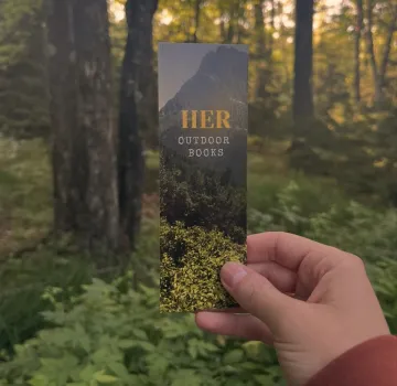Photo of a hand holding a Her Outdoor Books bookmark in a sunlit forest