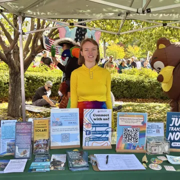 Anise, a young white woman with a bright yellow sweatshirt, stands behind a table under a canopy outdoors. The table has signage and materials related to the Sierra Club