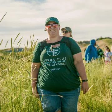 Kirsten, a white woman, walking toward the camera, smiling, with a green hat and shirt and sunglasses leading a hike in the Boise Foothills