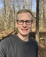 A man with short light brown hair and dark glasses stands with bare trees visibly behind him. He is smiling.