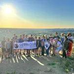 Picture of lots of people standing at top of a mountain overlooking a city holding a banner that says "Sierra Club OUTdoors Idaho" with the sun setting