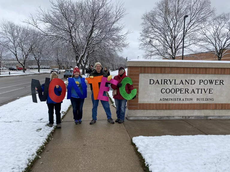Four Coulee Region Group members stand in front of Dairyland Power Cooperative's headquarters in La Crosse, holding up colored letters that say "NO NTEC".