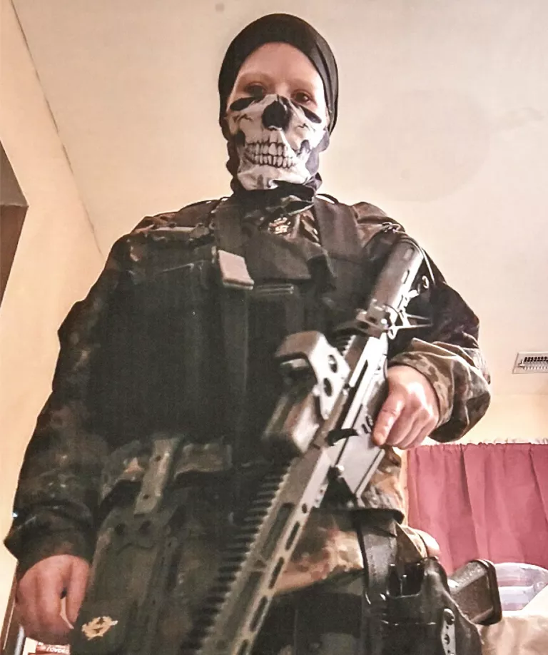 Sarah Beth Clendaniel wears camouflage clothing, a black head wrap, and a skeleton face mask. She's looking down at the camera and holding an assault rifle.