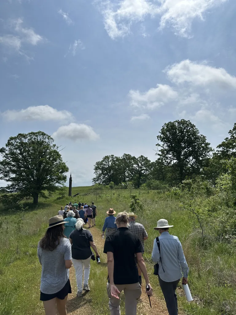 A group of hikers are walking away from the camera, on a path surrounded by green grass and tall trees at Eleven Point State Park.