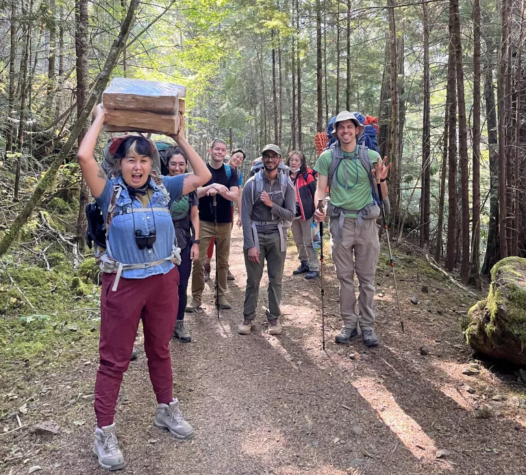 A diverse group of hikers on the trail, facing the camera with one hiker in front holder a bundle of campfire wood above them triumphantly