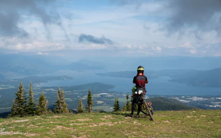 A bike leans against a person, who stairs out at the view from a mountain summit