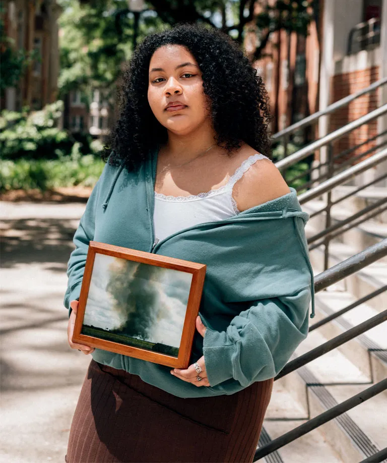 Madison Jones wears a blue hoodie and stands on concrete steps holding her framed image of sugarcane burning