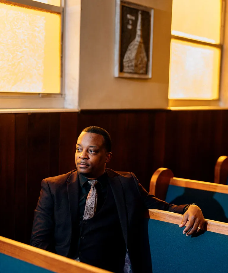 Reverend Messam wears a dark suit and tie and sits in a church pew