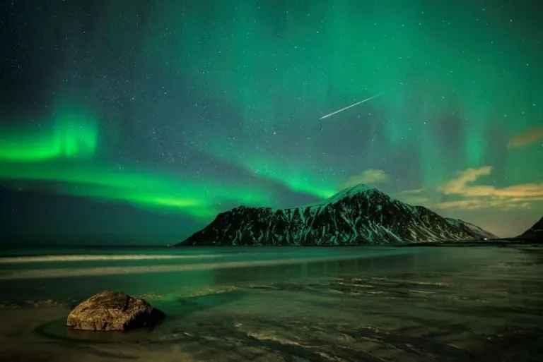 Aurora Borealis lights up the sky in green ribbons and a comet draws a white line across the sky. The foreground is a froze shore that reflects the green in the sky.