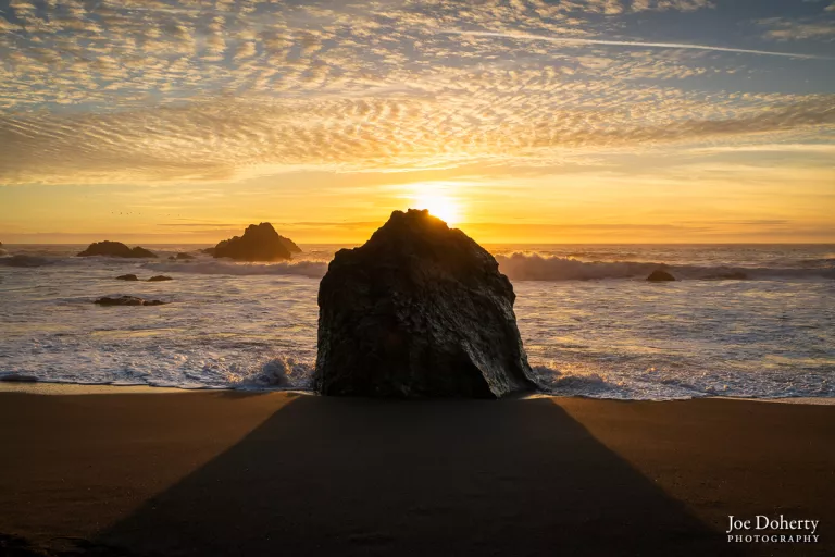 Wide sunset seascape large seastack in center forground casts a wide shadow with golden sky about the ocean horizon
