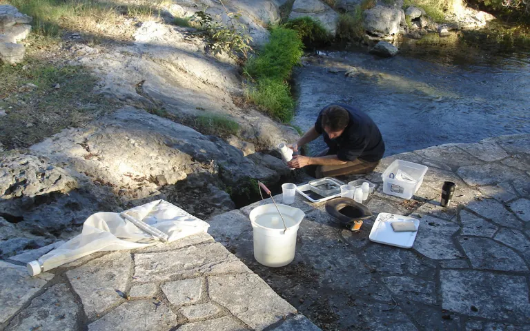 A man crouches in a rocky riverbed collecting samples. 