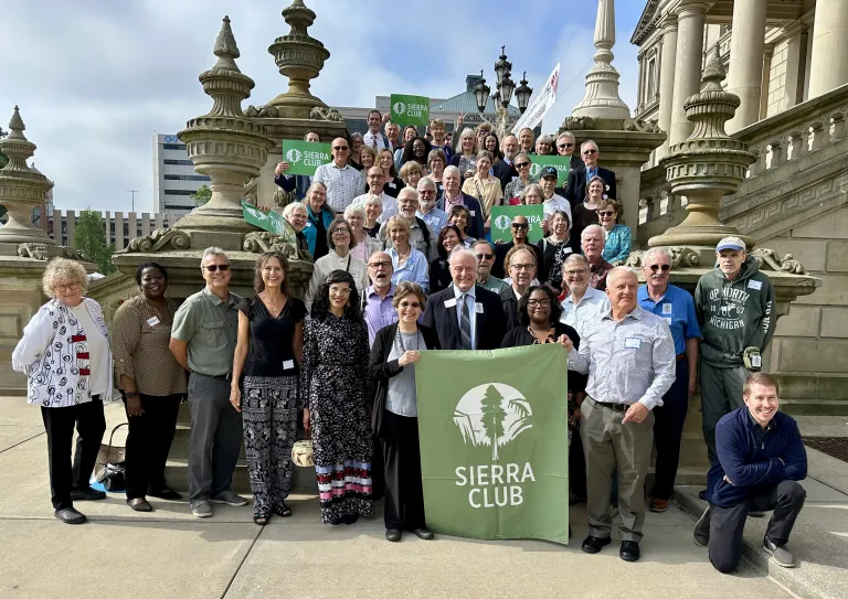 A diverse group of people are gathered on the steps of the state capitol in Lansing Michigan holding a Sierra Club Michigan Banner