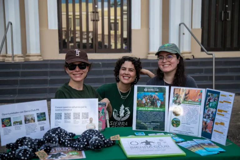 hree women stand behind an informational table for Sierra Club Puerto Rico, smiling and engaging with passersby. The table is covered with educational materials, flyers, and nature-themed displays promoting environmental awareness and community involvement.