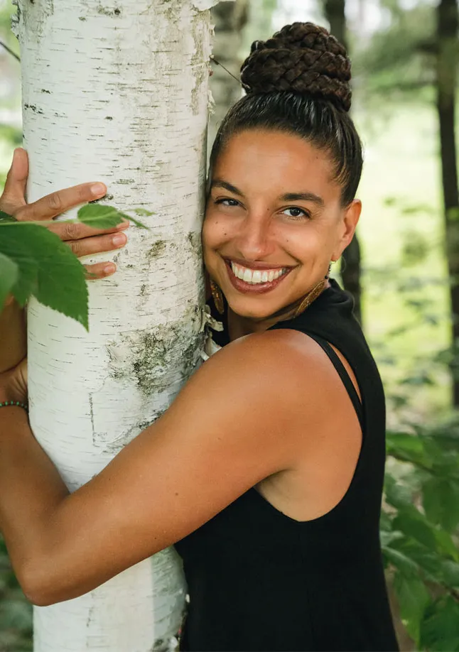 Leah Penniman wears a black top and smiles at the camera while hugging a white-barked tree.