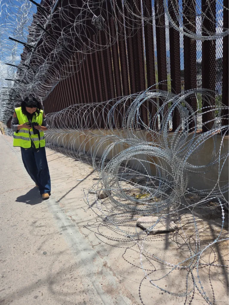 A person in a yellow vest stands by the border wall next to a pile of concertina wire